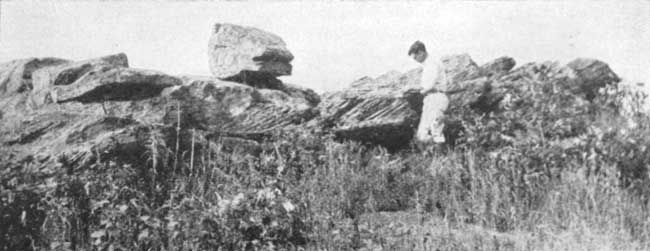 Black and white photo of researcher standing next to large squarish boulders, 2-5 feet thick, cross-bedding seen on sides of rocks.