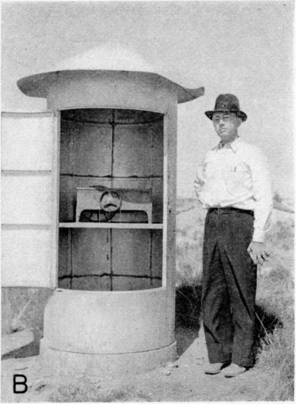 black and white photo of man standing next to covered well; cover is 6 feet tall and contains pulley to measure water levels
