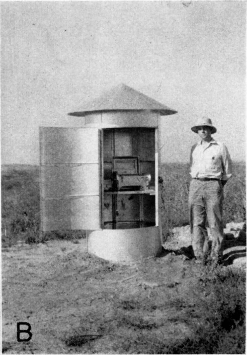 black and white photo of man standing next to covered well; cover is 6 feet tall and contains equipment to record water levels