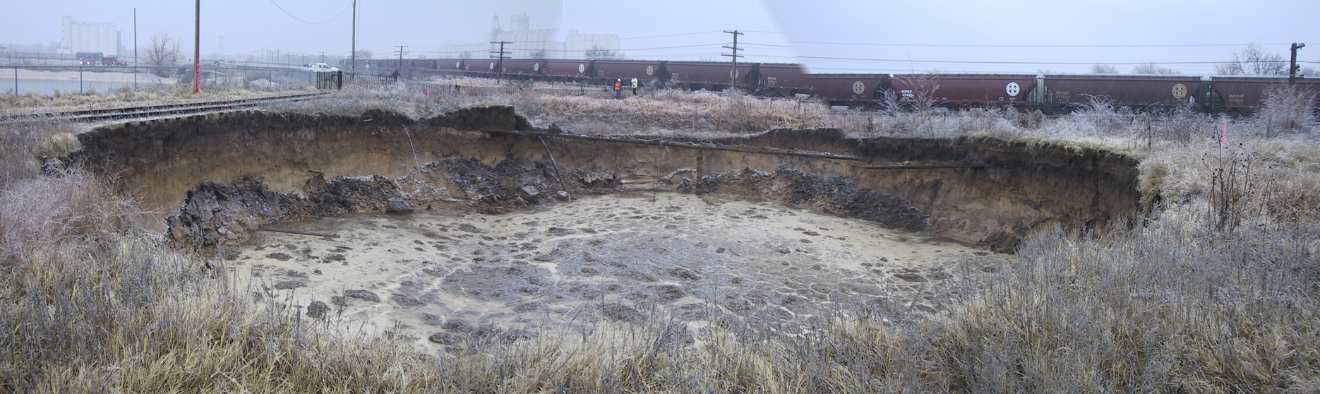 photo of sinkhole; about 115 feet in diameter and 20 feet deep; railroad tracks to left and behind hole are very close; train moving on far tracks