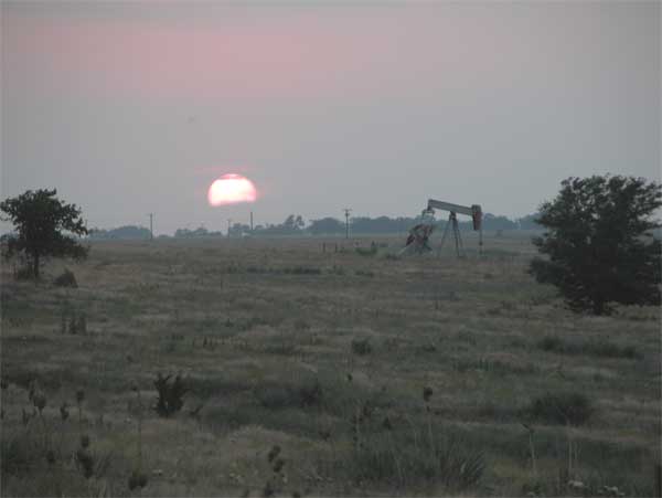 Evening view of grassy pasture; oil pumpjack in background.