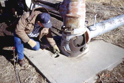 Photo shows worker lowering lead weight into hole to side of irrigation well.