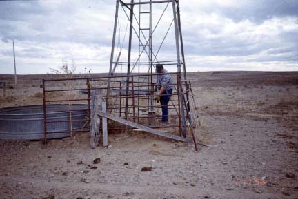Photo shows large metal stock tank with input pipe from windmill; only lower part of windmill tower is seen; crew member taking measurement.