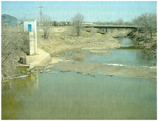 Color photo, river is low; winter photo as trees have no leaves; highway bridge in background; small cement dam in foreground