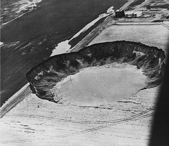 Black and white photograph, from air, showing water filled sinkhole covering corner of field; part of field road is missing.
