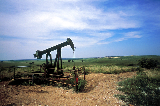 Pumpjack in small cleared area, top of small rise, green of Flint Hills in background, blue skies with light clouds