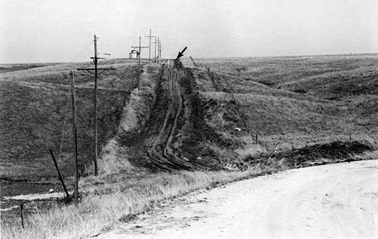 Black and white photo of unimproved road along line of power-telephone poles with Blue Hill Shale Member of the Carlile Shale at top of hill.