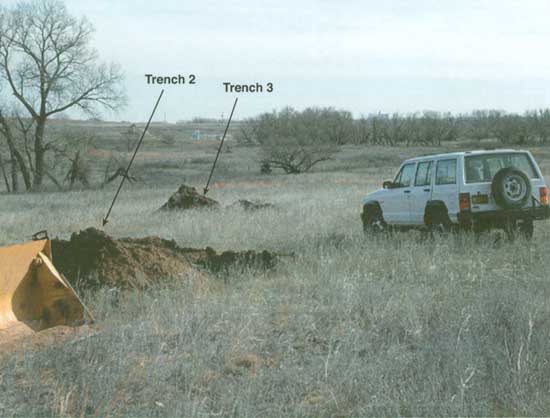 Photo of pasture; no snow, but trees and grasses dormant; two small piles of dirt show where trenches are; white survey vehicle to left.