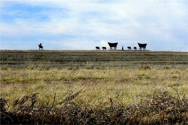 Photo of gentle hill in prairie; metal figures at top show a horse rider, a walker, and two wagons.