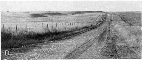 Black and white photo of dirt road in grasslands; rounded hills in background.