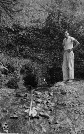 Black and white photo of man standing next to spring; water funneled into small pipe.