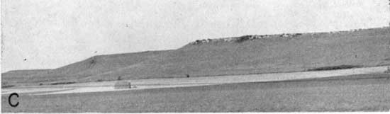 Black and white photo of isolated farm house in grassland, at base of gentle hill with limestone at top.