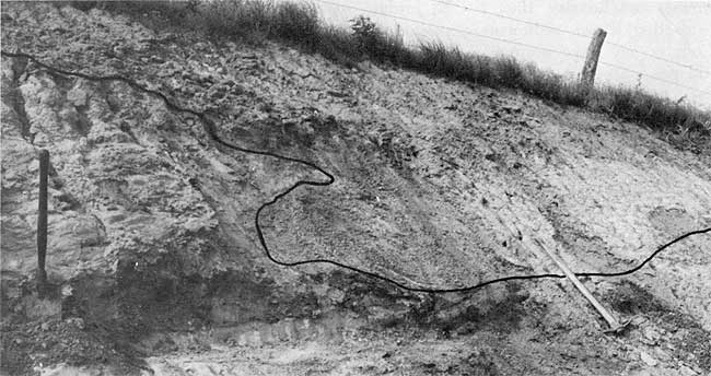 Black and white photo of roadcut; large miners pick for scale.