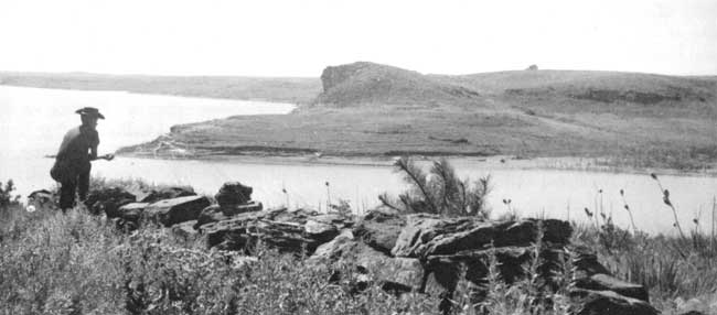 Black and white photo of kneeling figure overlooking lake; bluff with gentle slope on one side and sharp drop off on other is across lake.