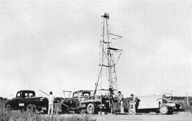 Black and white photo of portable drilling rig; looks to be 25-30 feet high; water tank in back; three men operating rig.