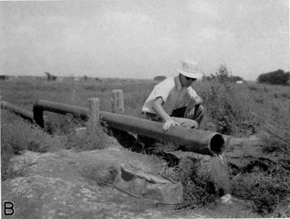 Black and white photo of man kneeling near 8-inch horizontal pipe, water flowing steadily out of pipe.