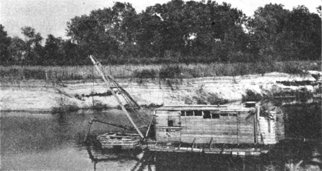 Black and white photo of small wooden boat in river; front of boat has crane holding a pipe into riverbed.