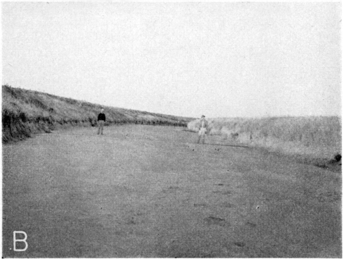 Black and white photo: two men in dry creek bed; very little relief; flat creek bed is 30-40 feet across.