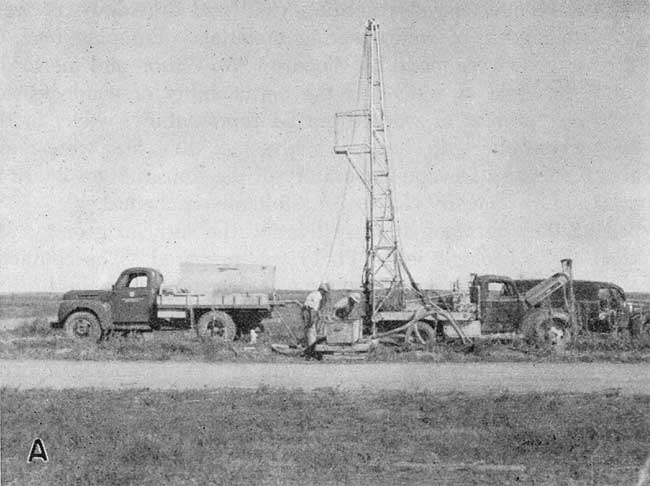 Small drilling rig on back of flat-bed truck drilling hole next to gravel road.