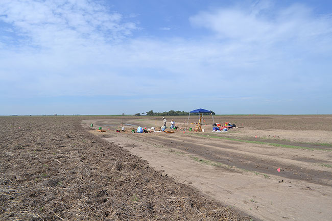 View of entire work site, blue sky with light clouds, dark blue shelter over workers digging.