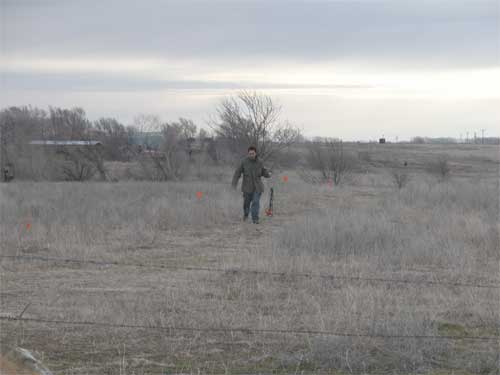 Technician walking through field; low, dormant grasses; barbwire fence in foreground; small trees and farm buildings in background; cold, overcast day