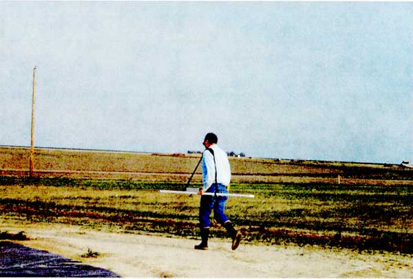 Researcher walking on dirt road carrying GEM2 unit.  Unit is a 6 foot or so flattish white pole, carried horizontally to ground.