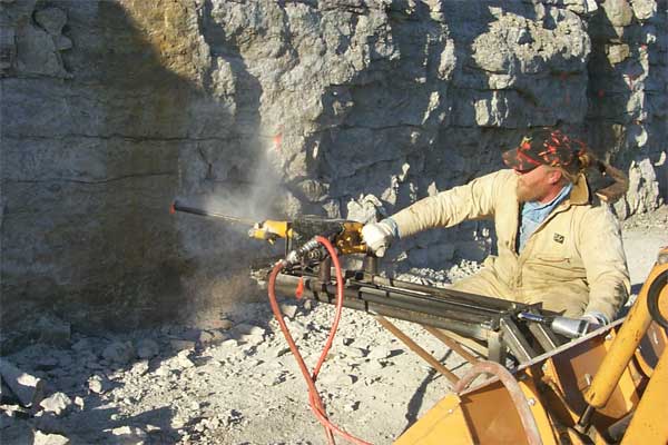 Color photo of hand-guided drill attached to front-end loader shovel.