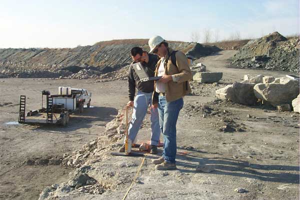Color photo; two people collecting data; one holds a laptop; the other holds GPR equipment; near face of quarry.