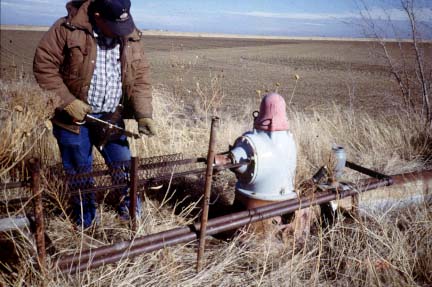 Irrigation well topped with pump (3-4 foot high); opening to side of unit; worker moving tape into hole.