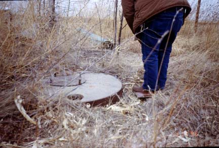 Well covered with plate, 3-3.5 feet across, just above ground level; surrounded by dried grasses.