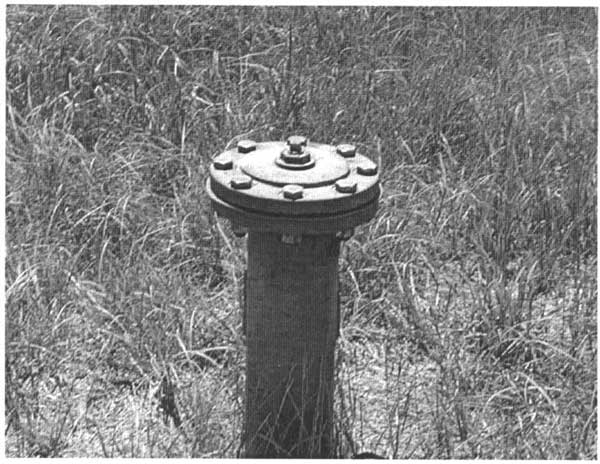 Black and white photo; well casing stands among short grasses.