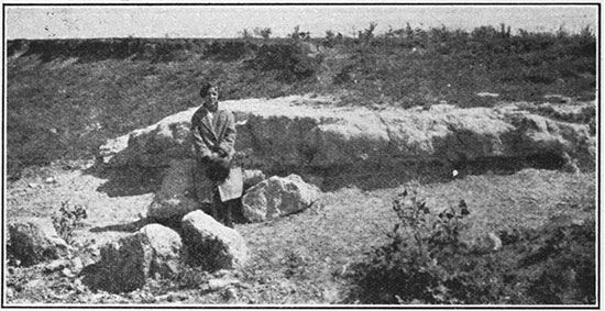 Black and white photo of volcanic ash outcrop; woman sitting on boulder for scale.