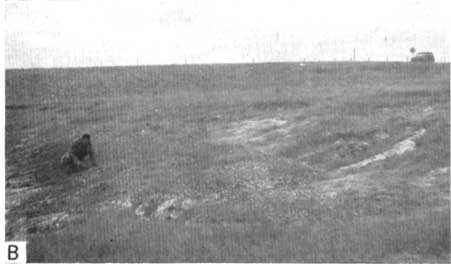 Black and white photo of kneeling man on very gentle slope; short grass covered with vehicle in background.