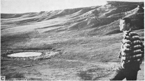 Black and white photo of man overlooking small water-filled depression; gentle hills in background.