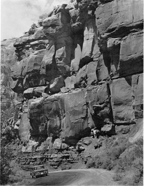 Black and white photo of car on narrow road below massive cliff of dark sandstone.