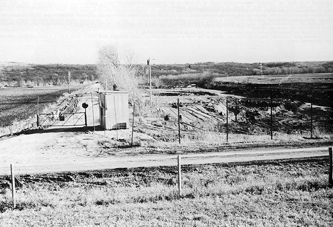 Photo of large fenced landfill, small line of hills in background; gravel road leading to gate with guard shack in center of photo.