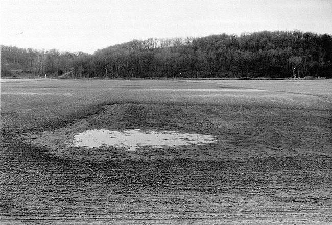 Field in winter after harvest, smoothed level with water-filled pond in forground; tree-covered hills in background.