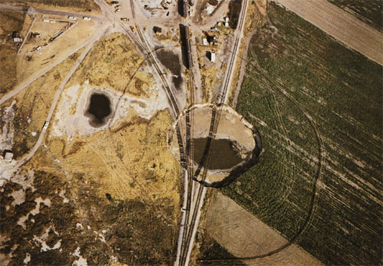 color photo of sink hole from above; very circular, easy to see trian tracks suspended above hole; water in center of hole and in gentler subsidenece feature to left. Photo courtesy Deming Studio, Hutchinson; was used on cover of book.