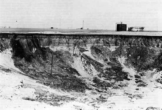 Ground-level photograph, black and white, showing sinkhole, oil tank in background; steep sides with rounded bottom; abandoned water well casing sticking up into air within hole.