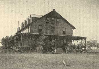 Hotel is a 2 1/2 story stone structure witha  wrap-around porch, small trees.  Small dog in front of photo.