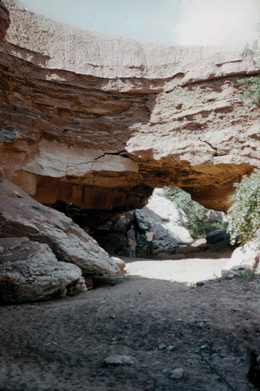Color photo of natural bridge; two people standing under it for scale.