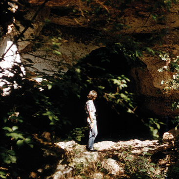 Color photo; person standing in front of small cave entrance.