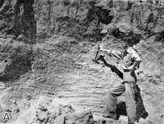 Black and white photo of man holding rock pick at gravel deposit.