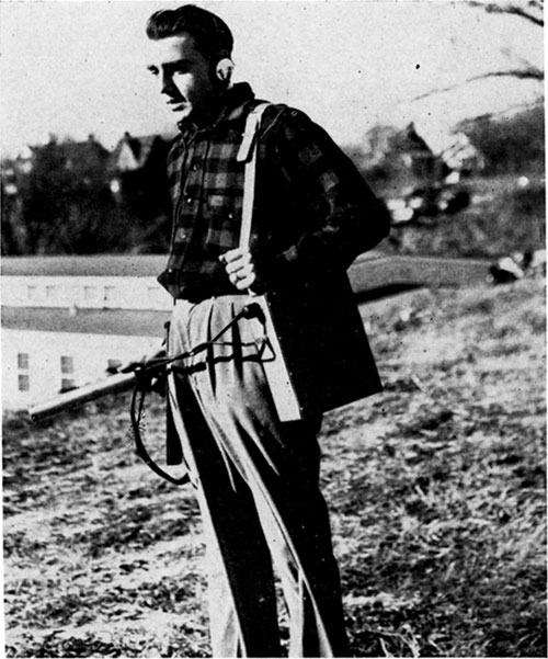 Black and white photo of man wearin Geiger-Muller counter; equipment hung over left shoulder with strap, foot-long cylindrical sensor in right hand.