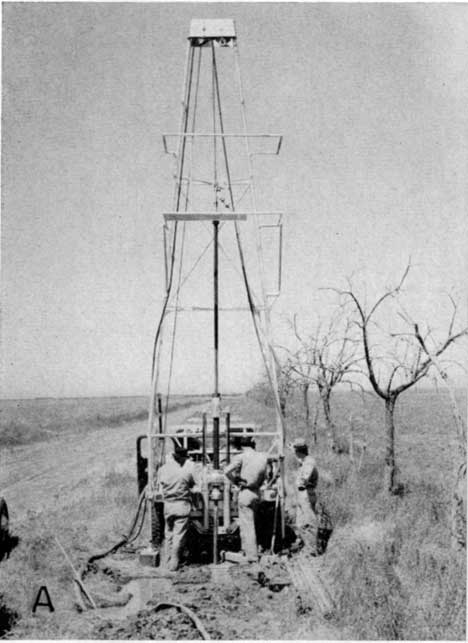 Black and white photo; three men around small drill rig; dirt or gravel road lined with small trees; no leaves on trees.