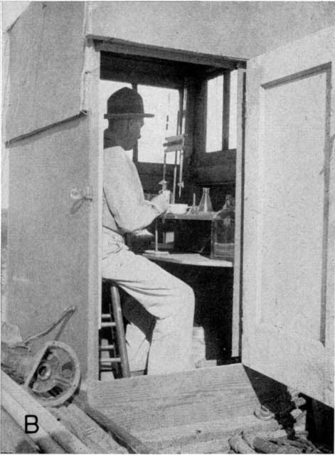 Black and white photo, sitting in portable lab; lab is wooden box, not much larger than he is, lab has small windows; looks like titration equipment in lab.