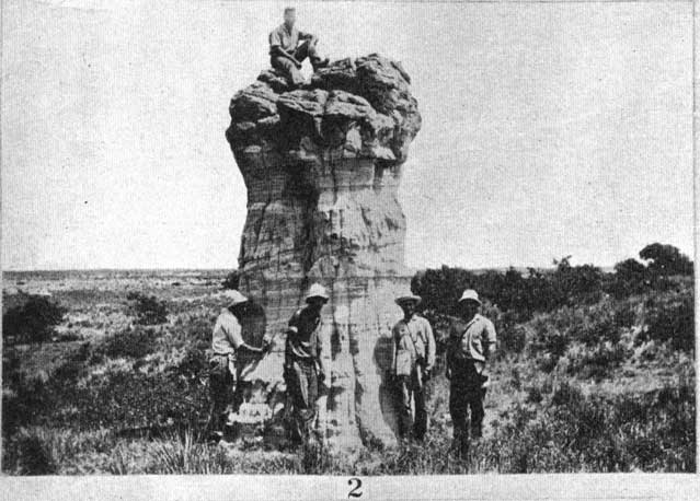 Black and white photo, small 'tower' of rock, 20-feet high and 8-10 feet in diameter; four men at base and one sitting on top