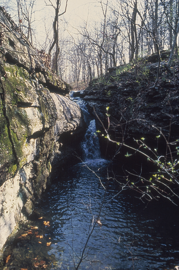 Narrow cleft of stone with waterfall at far end; trees have no leaves; waterfall is 10 feet or so high; rocks are light gray-tan in color, moss in small patches