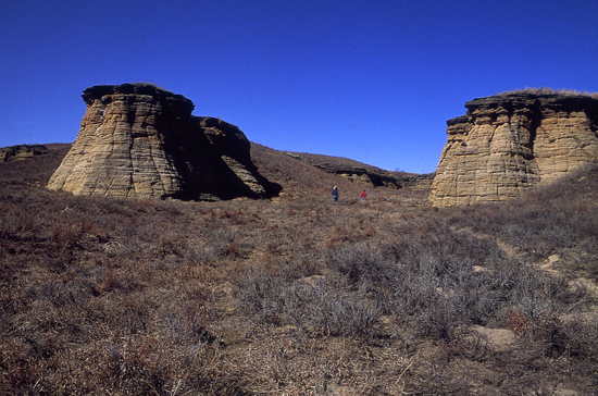 Steeply sided bluffs of tan-brown Dakota sandstone; vertical sides 30-40 feet high above brown grassy slopes; more resistant stone at top of bluffs adds a cap that extends out from cliff a foot or two