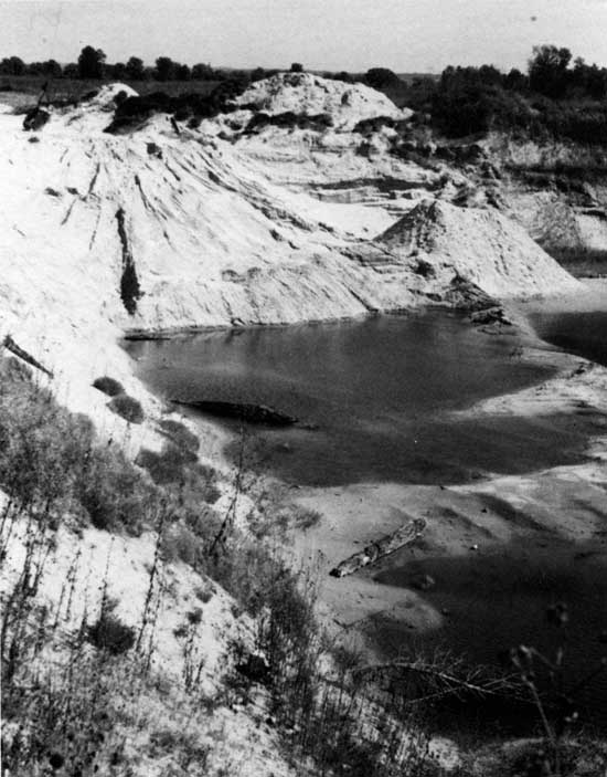 Black and white photo of abandoned sand pit; eroded sides, some brush, water in bottom of pit.